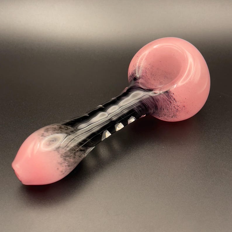 Op de afbeelding: Een roze en zwarte glazen pijp met een afgeronde kom en een lange, gebogen steel. De kom en het mondstuk zijn zachtroze van kleur, terwijl de steel een zwart-wit wervelontwerp heeft. De pijp is ontworpen om te roken.