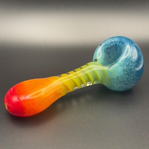 Op de afbeelding: Een kleurrijke glazen pijp met een kom en steel. De kom is blauw en turquoise, overgaand in geel en oranje langs de steel, eindigend in rood. De pijp heeft een gladde, afgeronde vorm met een getextureerde grip.