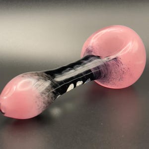 Op de afbeelding: Een roze en zwarte glazen pijp met een afgeronde kom en een lange, gebogen steel. De kom en het uiteinde van de steel zijn roze, terwijl de steel een zwart-wit wervelontwerp heeft. De pijp is ontworpen om te roken.