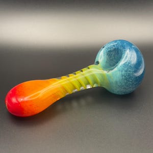 Op de afbeelding: Een kleurrijke glazen pijp met een blauwe kom en een steel die overgaat van geel naar oranje en rood. De pijp heeft een afgeronde kom en een lange, slanke steel met een textuurontwerp.