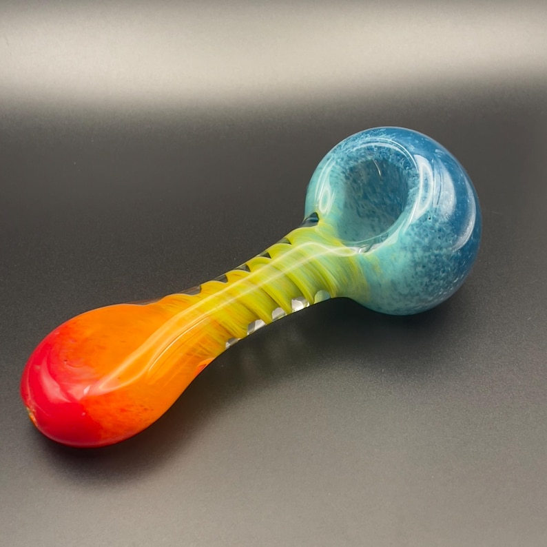 Op de afbeelding: Een kleurrijke glazen pijp met een blauwe kom en een steel die overloopt van geel naar oranje en rood. De pijp heeft een afgeronde kom en een lange, gebogen steel met een textuurontwerp. Het glas heeft een glanzende afwerking.