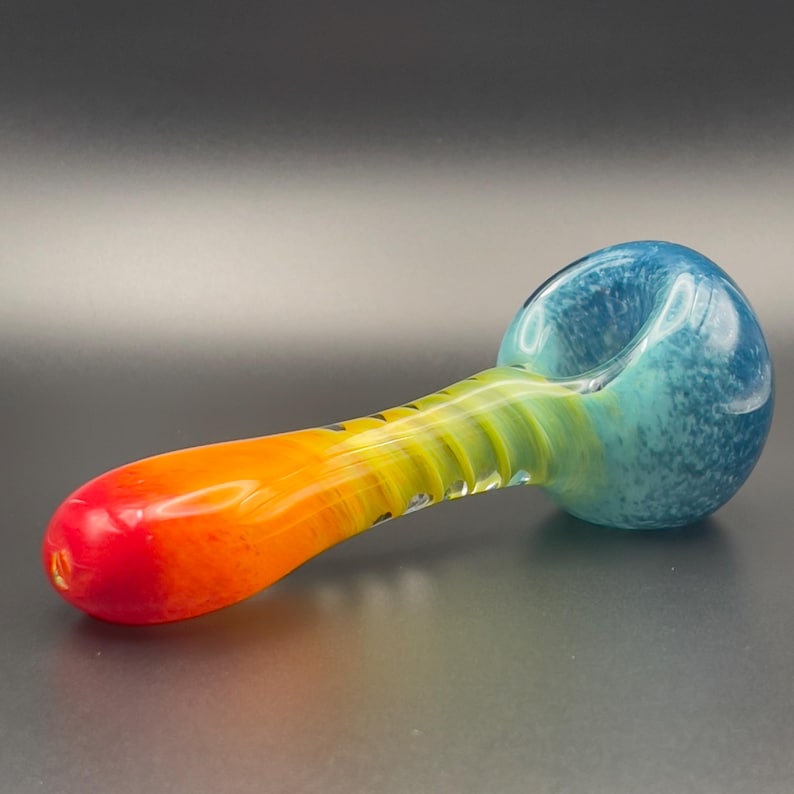 Op de afbeelding: Een kleurrijke glazen pijp met een kom en steel. De kom is een mix van blauw en turquoise, terwijl de steel overgaat van geel naar oranje en rood. De pijp is ontworpen om te roken.