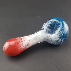 May include: A glass pipe with a red, white, and blue color scheme. The bowl is blue with white speckles, the stem is white with red speckles, and the mouthpiece is solid red. The pipe is designed for smoking.