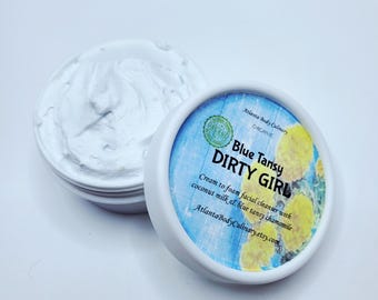 May include: A white jar of facial cleanser with the lid off, revealing a creamy, white product. The lid, with a blue and yellow design, reads "Blue Tansy DIRTY GIRL" and "Cream to foam facial cleanser with coconut milk & blue tansy chamomile."