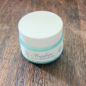 May include: A round, light blue glass jar of Magnifique Facial Moisturizer with a white screw-top lid. The label features a rose illustration and the text: Magnifique FACIAL MOISTURIZER, Organic/vegan/cruelty free, 40g. The jar is cylindrical.
