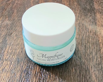 May include: A round, light blue glass jar of Magnifique Facial Moisturizer with a white screw-top lid. The label features a rose illustration and the text: Magnifique FACIAL MOISTURIZER, Organic/vegan/cruelty free, 1.4oz. The jar is cylindrical.