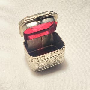 May include: A small, rectangular silver-toned metal box with an open hinged lid. The exterior features intricate embossed floral and leaf patterns. The interior of the lid is lined with a vibrant, translucent red material that catches the light.