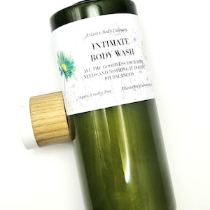 May include: A green bottle of "Intimate Body Wash" with a white label featuring text and a floral design. The bottle is held by a wooden and white holder. The label states "All the goodness your body needs and nothing it doesn't, pH balanced."