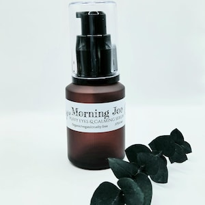 May include: A brown frosted glass bottle with a black pump dispenser and a clear cap. The white label reads Morning Joe Puffy Eyes & Calming Serum, Organic/vegan/cruelty free, .075/1.4oz. A dark green eucalyptus sprig is positioned next to the bottle.