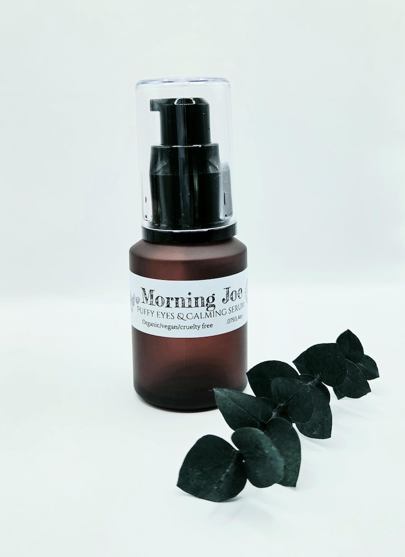 May include: A brown frosted glass bottle with a black pump dispenser and a clear cap. The white label reads Morning Joe Puffy Eyes & Calming Serum, Organic/vegan/cruelty free, .075/1.4oz. A dark green eucalyptus sprig is positioned next to the bottle.