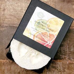 May include: A black box with a white label that says "Orange & Peppermint Loofah Foot Scrubbie". The label has a watercolor illustration of oranges and grapefruits. The box is open and a white loofah is visible.
