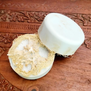 May include: A white, round soap bar sits atop a natural loofah sponge, with a portion of the sponge visible. The soap and sponge are on a carved wooden surface. The soap has a slightly aged appearance.