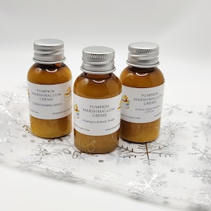 May include: Three small, amber-colored bottles with silver caps, each labeled "Pumpkin Marshmallow Creme." One is a conditioning rinse, another is shampoo and body wash, and the third is a 24-hour hydro moisture lotion. The bottles sit on a decorative, snowflake-patterned surface.