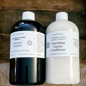May include: Two clear plastic bottles with black and white labels. The bottle on the left is black and labeled "Caffeine & Castor Formula Moisture Shampoo" and the bottle on the right is white and labeled "Caffeine & Castor Fallout Formula Nourishing Organic Conditioner". Both bottles are on a wooden surface.