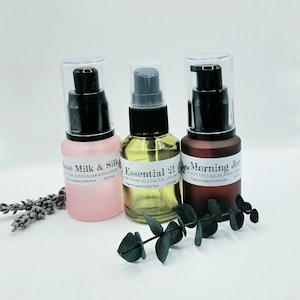May include: Three glass serum bottles with black pump dispensers. The left bottle is frosted pink labeled Rose Milk & Silk, the center is clear glass labeled Essential 21, and the right is frosted dark brown labeled Morning Joe. Each is 0.75 fl oz.