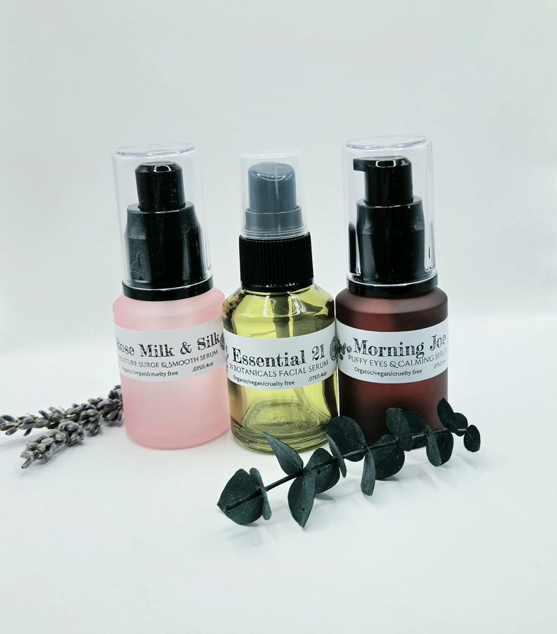 May include: Three glass serum bottles with black pump dispensers. The left bottle is frosted pink labeled Rose Milk & Silk, the center is clear glass labeled Essential 21, and the right is frosted dark brown labeled Morning Joe. Each is 0.75 fl oz.
