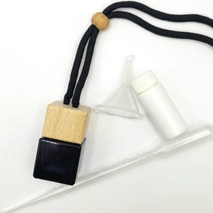 May include: An air freshener with a black cord and wooden bead. The air freshener has a natural wood block on top of a black square. Also included are a small funnel, a white bottle, and a clear dropper.