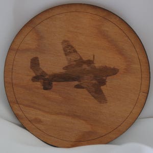 May include: A wooden coaster with a silhouette of a vintage airplane burned into the surface. The coaster is circular and has a dark brown wood grain.