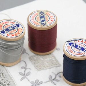 May include: Three spools of thread with wooden bobbins. The spools are labeled "Clark's O.N.T. Button & Carpet" and feature red, blue, and gray thread.