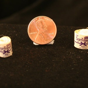 May include: Two miniature rolls of ScotTissue facial tissue, each with a purple and white label, next to a US penny.