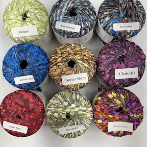 Peut inclure: Neuf écheveaux de fil coloré sont disposés en grille. Les couleurs de fil comprennent le vert, le bleu, le marron, le rouge, l'or et le violet. Chaque écheveau porte une étiquette avec un nom de couleur, comme "Peridot" et "Bright Rose."