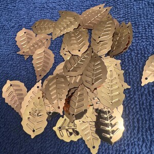May include: A collection of metallic gold-colored leaf-shaped embellishments. Each leaf has detailed vein patterns and a small hole at the top. The leaves are scattered on a blue textured surface.