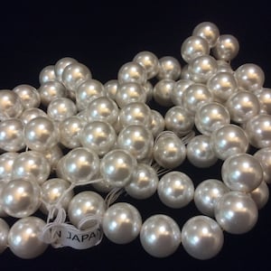 May include: A strand of white faux pearls, strung together on a white thread. The pearls are round and smooth, with a slight iridescent sheen. The strand is labeled "Made in Japan".