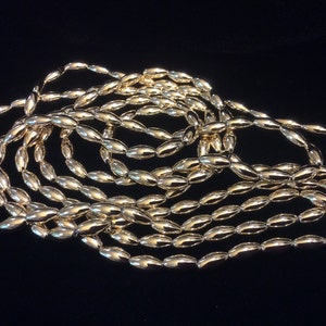 May include: A strand of gold-colored oval beads. The beads are smooth and shiny, and they are arranged in a loose pile.