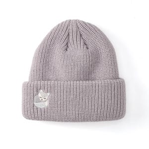 May include: A light gray knit beanie with a folded brim. The beanie features a small embroidered fox design on the front. The fox is depicted in shades of gray and white, with closed eyes, suggesting a sleeping or relaxed pose. The hat is made of a ribbed knit material.