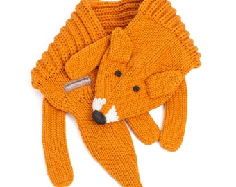 Op de afbeelding: Een gebreide oranje sjaal in de vorm van een vos, met een vossenkop met een witte snuit, een zwarte neus en twee oren. De sjaal heeft een geribbelde textuur en bevat een klein grijs stoffen label met de tekst Peppercorn kids erop gestikt.