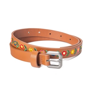 May include: A tan leather belt with colorful embroidered flowers. The belt features a silver-toned buckle and is designed with a floral pattern along the top edge. The belt is a thin style, suitable for various outfits.