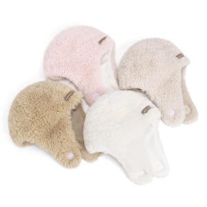 May include: Four fuzzy earflap hats in shades of light pink, beige, tan, and cream. Each hat features a soft, textured exterior, a smooth interior lining, and a small rectangular brand tag on the side. The earflaps include small circular fasteners.