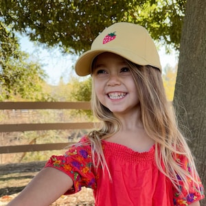 May include: A young girl with long blonde hair smiles while wearing a yellow baseball cap with a red strawberry embroidered on the front. She is wearing a red off-the-shoulder top with colourful floral embroidery on the sleeves.
