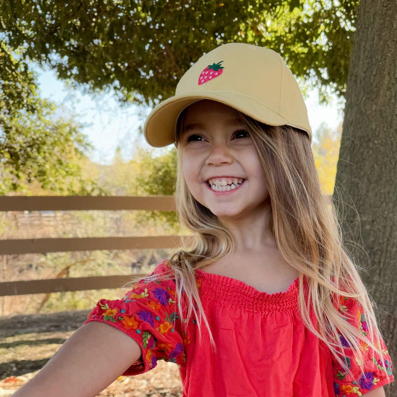 May include: A young girl with long blonde hair smiles while wearing a yellow baseball cap with a red strawberry embroidered on the front. She is wearing a red off-the-shoulder top with colourful floral embroidery on the sleeves.