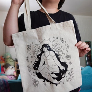 Puede incluir: Una bolsa de tela blanca con un dibujo de l&iacute;nea negra de una mujer con alas y un halo. La mujer lleva un vestido y tiene el pelo largo. El fondo est&aacute; lleno de flores y estrellas.