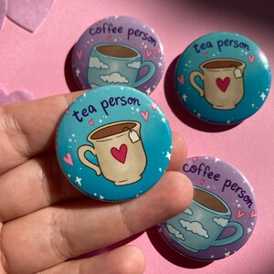 May include: Four circular badges with coffee and tea cup designs. Two badges say "coffee person" and show a cup with clouds. Two badges say "tea person" and show a cup with a heart. The background is pink.