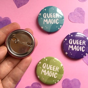 Puede incluir: Cuatro insignias redondas con el texto "QUEER MAGIC" en escritura blanca. Las insignias son de color verde azulado, morado y verde con un diseño de remolino o estrella. Una insignia se muestra desde la parte posterior, con un cierre de alfiler de seguridad. El fondo es rosa con confeti de corazón morado.