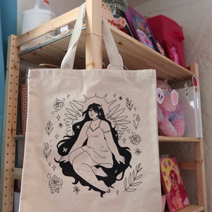 Puede incluir: Una bolsa de tela beige con una ilustraci&oacute;n en blanco y negro de una mujer con alas y un halo. La mujer est&aacute; rodeada de flores y estrellas. El texto "So. Many. Stars." est&aacute; impreso en la bolsa.
