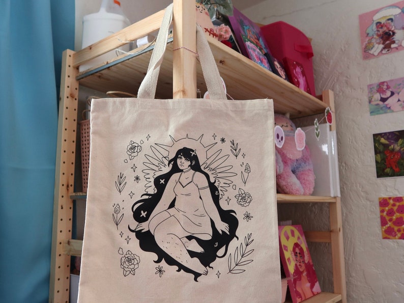 Puede incluir: Una bolsa de tela beige con una ilustraci&oacute;n en blanco y negro de una mujer con alas y un halo. La mujer est&aacute; rodeada de flores y estrellas. El texto "So. Many. Stars." est&aacute; impreso en la bolsa.