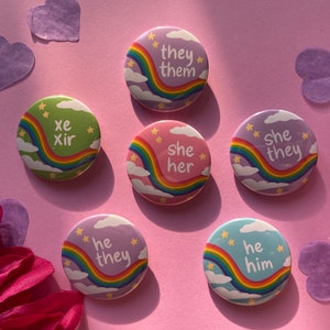 Many pins on a pink background with heart confetti and flowers at the sides. The pins are all pastel colors with a rainbow stripe, stars, and clouds, and have different pronouns written in lowercase white text.