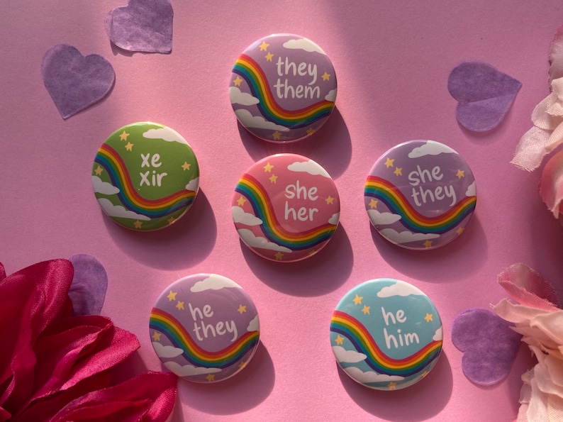 Many pins on a pink background with heart confetti and flowers at the sides. The pins are all pastel colors with a rainbow stripe, stars, and clouds, and have different pronouns written in lowercase white text.