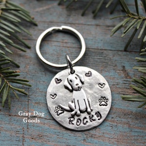 Peut inclure: Une médaille pour chien en argent avec une illustration de chien, des cœurs et des empreintes de pattes. Le nom "Rocky" est estampillé en dessous. La médaille est attachée à un porte-clés. Le texte "Gray Dog Goods" est imprimé.