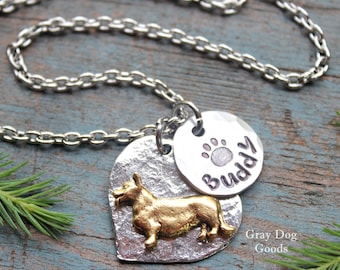 May include: Silver chain necklace with a heart-shaped pendant featuring a gold-coloured dog silhouette and a round pendant with the word "Buddy" and a paw print.