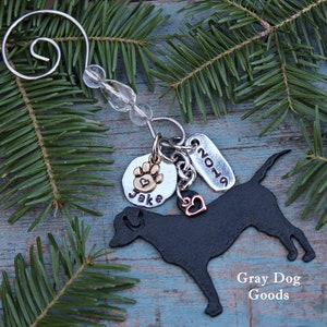 May include: A black metal dog silhouette ornament with a paw print charm, a heart charm, and a tag that reads "2019". The ornament is attached to a silver wire with clear beads. The text "Jake" is on the paw print charm. The words "Gray Dog Goods" are in the image.