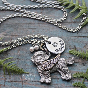 May include: Silver chain necklace with a silver dog charm with wings, a silver charm with a paw print and the word "ROCKY" on it, and two white pearl beads. The necklace is on a blue wooden surface with green fern leaves.