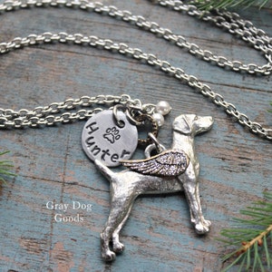 May include: Silver necklace featuring a silver dog charm with wings, a silver charm engraved with a paw print and the word "Hunter", and two white pearl beads. The necklace is displayed on a wooden surface with green pine branches.