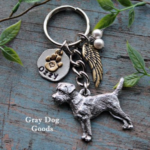 May include: Silver charm keychain featuring a small dog figurine, a paw print charm with the name "Ozzy" and a gold angel wing charm with two white pearl beads. The keychain is on a wooden surface.