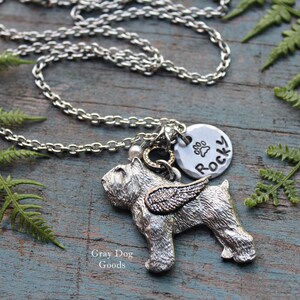 Peut inclure: Collier en argent avec un pendentif représentant un chien avec des ailes, un pendentif rond avec une empreinte de patte et le mot "Rocky" inscrit dessus, ainsi qu'une petite perle dorée. Le collier est posé sur une surface en bois avec des fougères vertes.