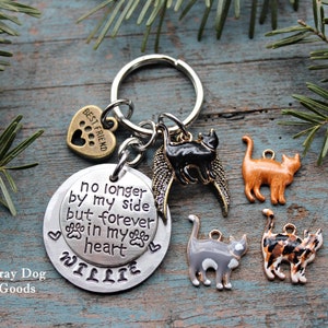 May include: A silver keychain with a round charm that reads "no longer by my side but forever in my heart" and the name "WILLIE". The keychain also features a small heart charm with the words "Best Friend" and paw prints, a black cat charm with wings, and three other cat charms in orange, gray, and black and white.