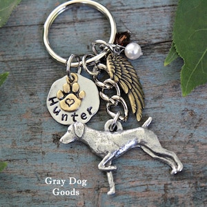 May include: A silver and gold charm keyring with a dog paw print, a heart, and the name "Hunter" engraved on it. The keyring also features a silver dog charm, a silver angel wing charm, and a pearl. The keyring is on a wooden surface with green leaves.